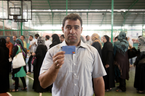 Syrian refugee Ali Ahmad Farhat holds his MasterCard / WFP e-card as others stand in line to receive theirs, at a basketball court. (Photo: Business Wire)