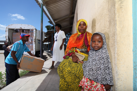 タンザニアのダルエスサラーム県北部ゾーン地域の診療所の外で、トラックが診療所に直接医薬品を届ける中、待っている母親のナスラ・アリーさんと子供。親子は、プロジェクト・ラストマイルの実施前には簡単に利用できなかった治療を受けようと待っている。（写真：ビジネスワイヤ）