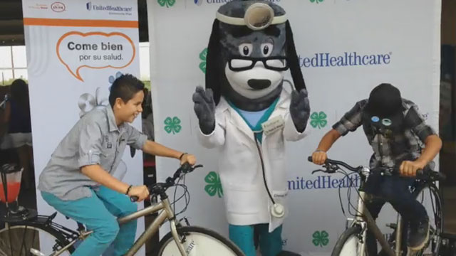 (L-R) Jovannie Rancheria (11) and Aaron Cruz (13) from Pategonia Montessori School are cheered on at the Santa Cruz County Fair in Sonoita by UnitedHealthcare mascot Dr. Health E. Hound and fellow students, as they make their own healthy smoothies on new blender bikes donated to Arizona 4-H as part of a $40,000 grant from UnitedHealthcare (Video: Espe Greenwood).