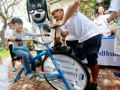 Sarah, left, pedals a specially made smoothie bike today at the annual Olivewood Gardens and Learning Center Day of Play in National City, Calif., while UnitedHealthcare mascot Dr. Health E. Hound and employee volunteer Jose Gomez cheer her on. The specially made smoothie bikes are designed to help kids and their families connect an active lifestyle with healthy eating. (Photo credit: Sandy Huffaker)
