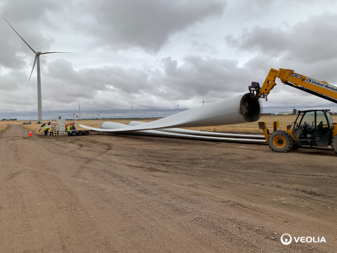 A wind turbine blade being prepared for removal after it has reached the end of its life cycle, with a nearby wind turbine in the backdrop. Blades like these are now being repurposed by Veolia North America after reaching the end of their life cycle, which typically lasts about 20 years. (Photo: Business Wire)