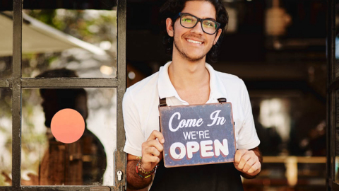 Male small business owner holding a sign that says “Come in we’re open.” (Photo: Wells Fargo)