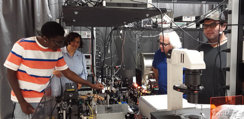 Experimental setup for a magnetometer using alkali sodium atoms in Tripathi's DSU lab. The yellow glow from a sodium laser is visible in the picture. Left to right: Lawrence Taylor, a freshman pursuing a BS in Engineering Physics; Tripathi; Robin Depto, a BS Electrical Engineering student at Delaware Technical Community College; and Mauricio Pulido, who is pursuing an MS in Applied Optics. (Photo: Business Wire)