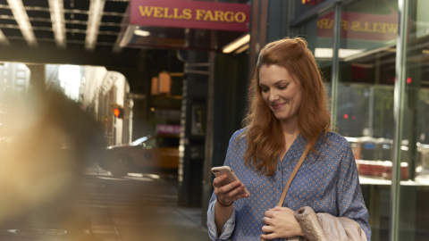 A woman looks at her phone with Wells Fargo sign in background. (Photo: Wells Fargo)