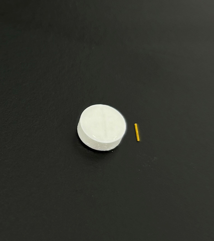 inflammasome's sustained release implant contains the first of a new class of inflammasome inhibitor drugs, Kamuvudines, also developed by the company. The new drug is shown here next to a standard aspirin tablet. Following implantation, tiny amounts of  release Kamuvudine will be released at a consistent rate directly to the back of the eye for an initial period of three months during  the upcoming Phase 1/2 clinical trial. (Photo: Business Wire)