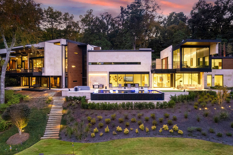 Shown here: the rear exterior of the residence glows in the evening. The resort-style outdoor living area features a heated deck in Ipe wood, multiple lounge areas, and an onyx pebble infinity-edge pool with integrated spa. Steps lead down to the manicured lawn and lake, where there is a lakeside firepit lounge, Ipe wood boathouse and dock (not pictured). GeorgiaLuxuryAuction.com. (Photo: Business Wire)
