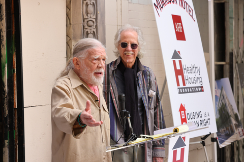Henry Diltz (Left) addresses the crowd during a ceremony celebrating AHF’s acquisition of the historic Morrison Hotel, a 1914 Single Room Occupancy (SRO) building in downtown Los Angeles as John Densmore, drummer for The Doors, looks on Tuesday, December 19, 2023. The Doors and photographer Henry Diltz made the Morrison famous with the band’s release of its landmark 1970 album featuring Diltz’s iconic cover photo. AHF purchased the gutted building for its Healthy Housing Foundation, saving the Morrison from demolition and, via adaptive reuse, will now convert it into 111 units of low-income housing for people in need.(Photo: Business Wire)