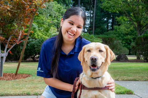 A young woman with long dark hair smiles while sitting with a yellow Labrador Retriever from Guide Dogs for the Blind. (Photo: Business Wire)