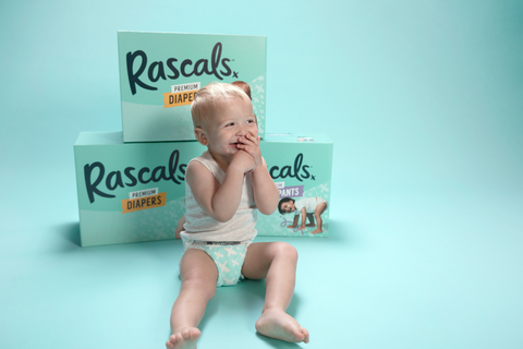 A baby sitting happily in front of three Rascals Premium Diaper boxes, showcasing the brand's packaging. (Photo: Business Wire)