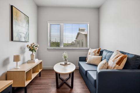 original A cozy, modern sitting area featuring a blue sofa with neutral-toned pillows, a wooden coffee table with a small plant and book, and a console table with a lamp and flowers. A large abstract painting hangs above, and natural light filters through white blinds, creating a warm, welcoming atmosphere.