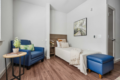 thumbnail A serene, thoughtfully furnished bedroom featuring a single bed with a wooden headboard, layered pillows, and a soft blanket. A blue armchair with a green cushion sits beside a small round table holding books and a potted plant. A nightstand with fresh flowers adds a gentle touch, while a framed nature scene above the bed and natural wood flooring create a peaceful, welcoming atmosphere.