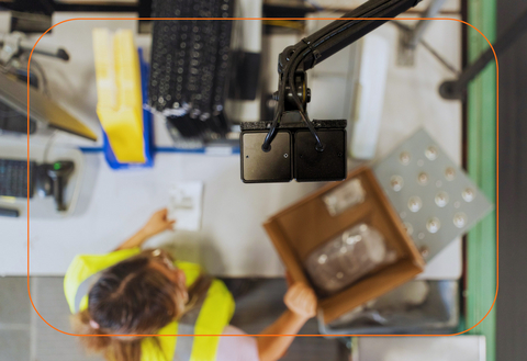 Overhead view of a Rabot Vision AI camera at a pack station inside a Yusen Logistics (Americas) warehouse. The orange overlay shows the camera's AI field of view monitoring the station surface as a worker packs items, providing real-time guidance and quality verification.