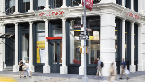 Corner view of a Wells Fargo branch at a busy city intersection with pedestrians and street signs.