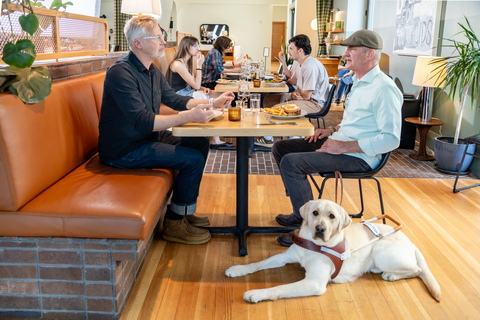 original Friends enjoy a meal; a guide dog lies at man's feet. Photo demonstrates the importance of access rights for people with guide dogs.