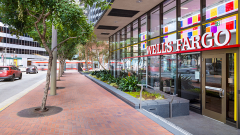 Exterior view of a Wells Fargo branch along a city sidewalk with trees and passing traffic.